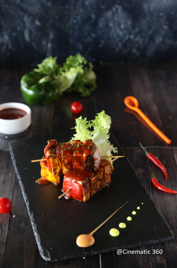 Gourmet grilled paneer tikka skewers with sauce glaze served on a black slate board with fine-dining sauce plating by Cinematic360.