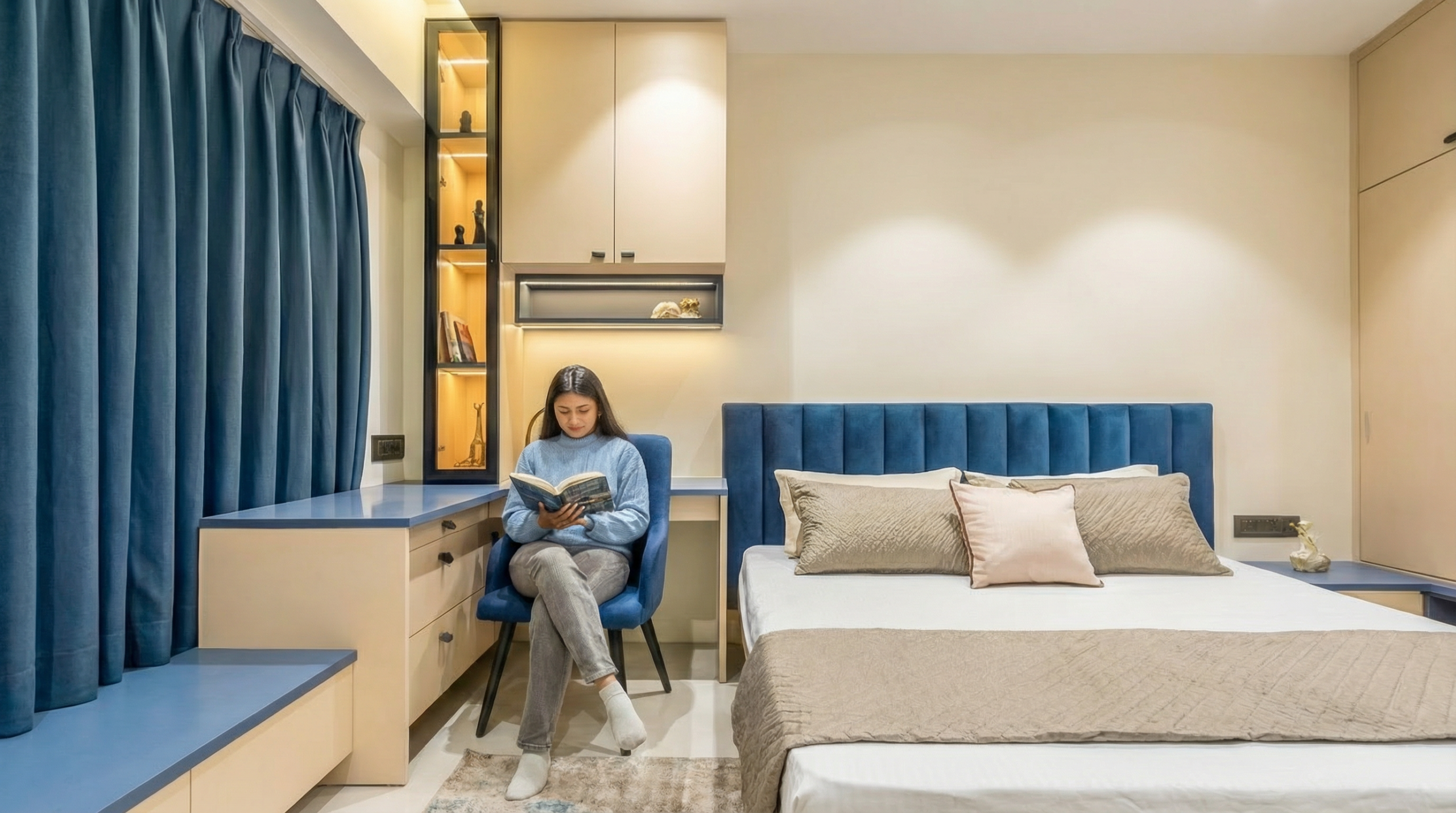 Blue and beige bedroom study corner with lighted bookshelf and curtains.