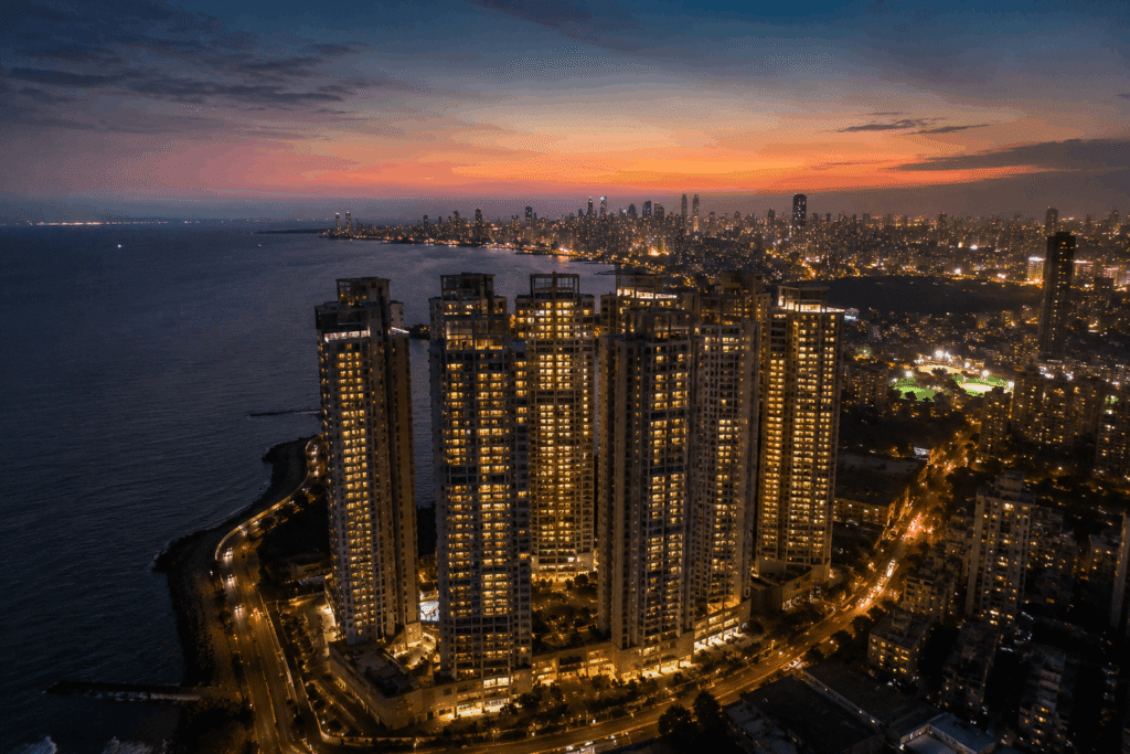 Aerial drone shot of luxury seaside high-rise apartments in Mumbai at sunset, showcasing city lights and the coastline, captured by Cinematic 360 premium drone photography services.