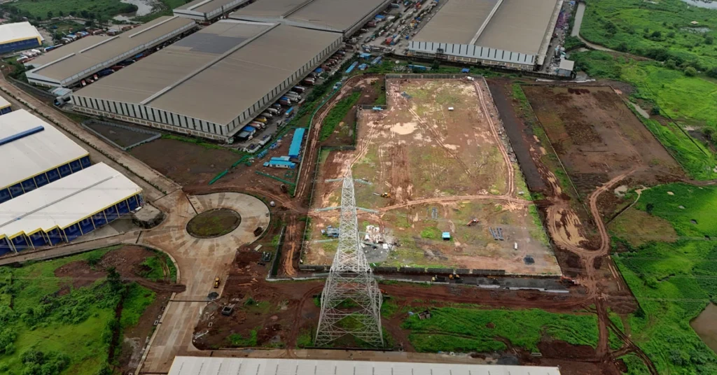 Aerial drone photograph of an under-construction industrial land plot and warehouse site tracking in Bhiwandi, Mumbai, showing cleared muddy ground, a prominent power transmission tower, surrounding logistics parks, and connectivity roads for a developer's launch blueprint.