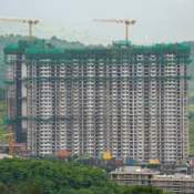 An aerial drone shot of a massive high-rise residential building under construction with active tower cranes against a lush green hillside backdrop, documenting project timeline progress tracking by Cinematic360.