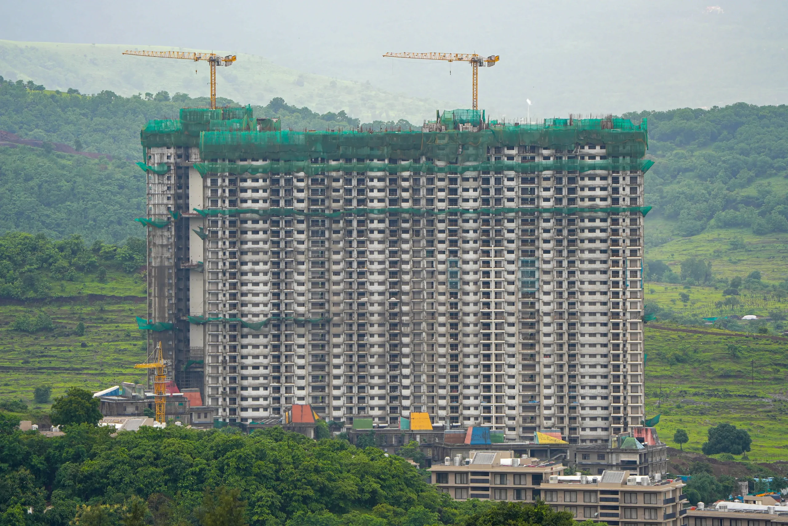 An aerial drone shot of a massive high-rise residential building under construction with active tower cranes against a lush green hillside backdrop, documenting project timeline progress tracking by Cinematic360.