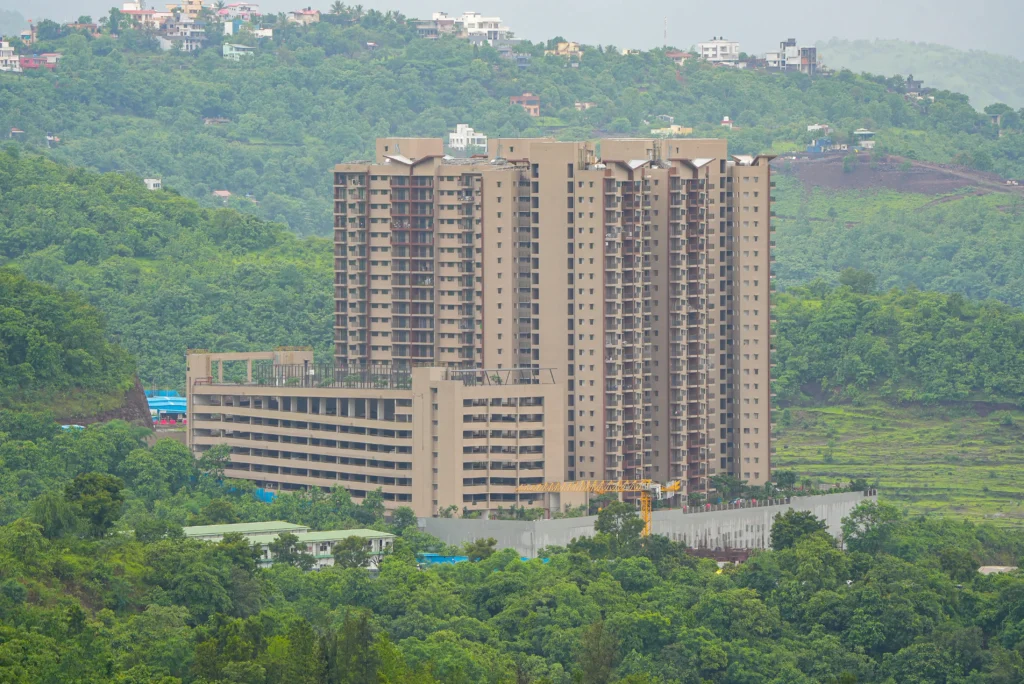 A high-angle aerial drone photograph of a massive completed luxury high-rise apartment complex featuring an integrated multi-level parking structure, situated against a dramatic lush green hillside backdrop by Cinematic360.