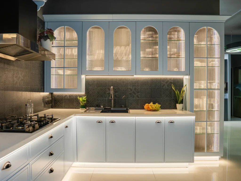 Professional interior photograph of a modern white modular kitchen with arched glass cabinets and warm LED lighting