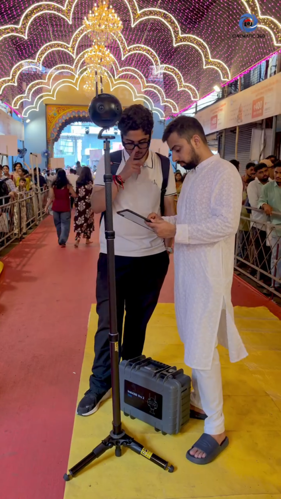 o Cinematic360 crew members reviewing live preview on tablet next to Insta360 Pro 2 on carbon fiber monopod inside decorated festival corridor Mumbai