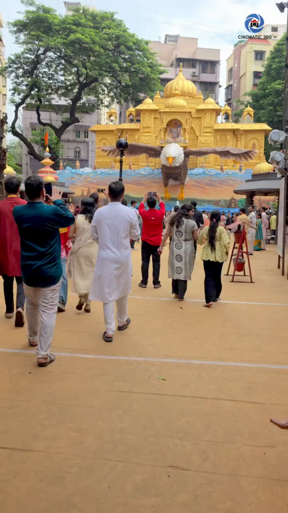 Insta360 Pro 2 on slim monopod positioned in front of elaborate Lalbaug Ganesh festival golden pandal with eagle sculpture and visitors walking toward it